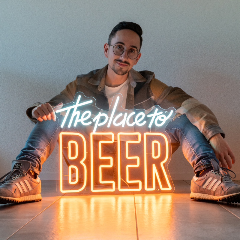Homme assis tenant une enseigne néon personnalisée « The place to BEER » lumineuse orange et blanche