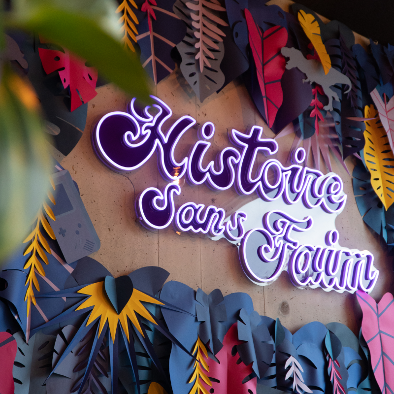 Logo néon violet « Histoire Sans Faim » sur mur décoratif coloré de restaurant